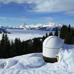 A snow-covered observatory amid a pine forest overlooks a vast, cloud-filled valley with distant snow-capped mountain peaks under a clear blue sky.