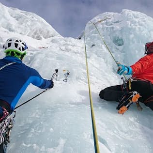 Two climbers in colorful gear ascending a steep icy slope with ice axes and ropes.