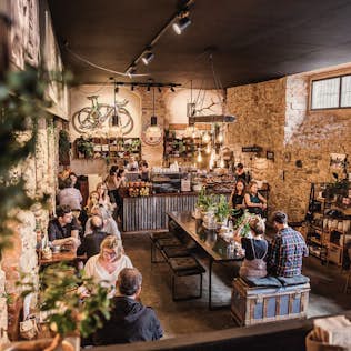 Interior of a rustic café filled with customers, featuring exposed stone walls and modern décor, with bicycles hanging and plants scattered around.