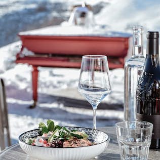 Outdoor dining setting with a meal and wine on a table, overlooking a snowy mountain landscape.