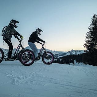 Two cyclists on fat tire bikes pause on a snowy mountain trail at dusk, with a colorful sunset and pine trees in the background.