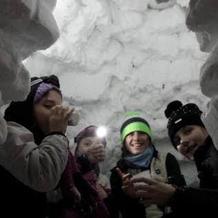 Four children smiling inside a snow fort, with one child drinking from a thermos, illuminated by soft light.