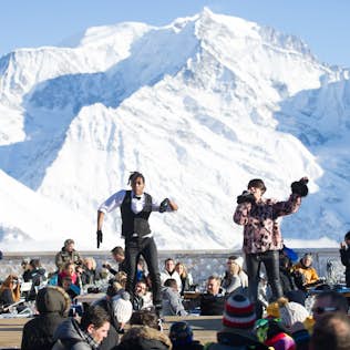 Two performers dancing on an outdoor stage at a snowy mountain resort, with an audience in winter attire watching them and a large, snowy mountain peak in the background.
