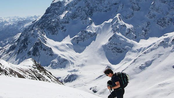 A person skis uphill on a snowy mountain slope, with a large mountain range in the background under a clear blue sky.