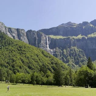 A panoramic view of a lush green valley with a single person walking across it, set against towering jagged mountains under a clear blue sky.