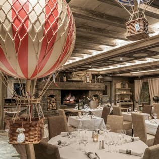 Rustic restaurant interior with wooden beams, fireplace, and hot air balloon decorations hanging from the ceiling. tables are set with white linen and fine dinnerware.