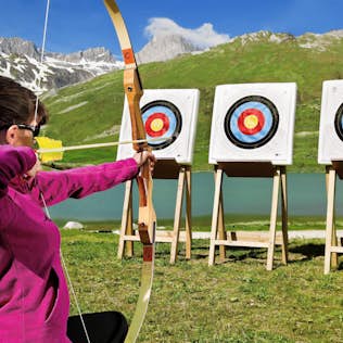 A woman in a pink jacket practicing archery, aiming at a target in the scenic mountainous and lake area of Tignes under a clear sky.