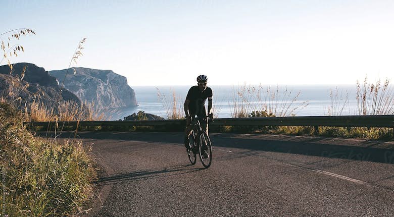 Cyclist in black attire riding on a coastal road with ocean and cliffs in the background during sunset.