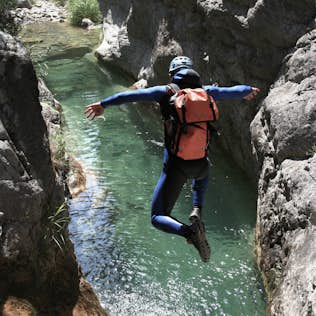 A person in a wetsuit, helmet, and life jacket jumps into a narrow canyon river with arms outstretched.
