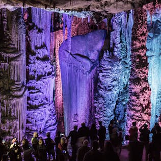 Visitors observing a large, illuminated stalactite formation inside a cave with colorful lighting.