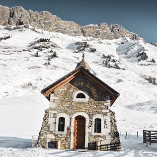 A small stone chapel with a red door, set against a snowy mountain landscape under a clear sky.