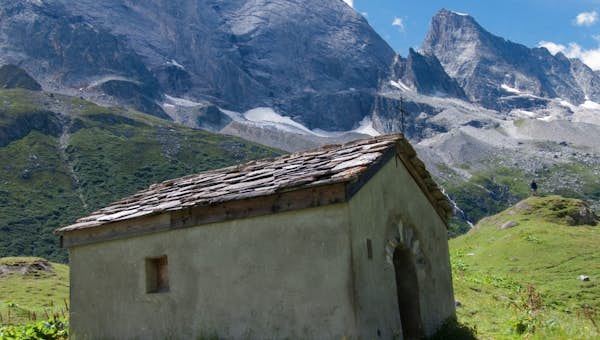 A small stone hut stands on a grassy hillside with rocky mountains in the background under a blue sky.
