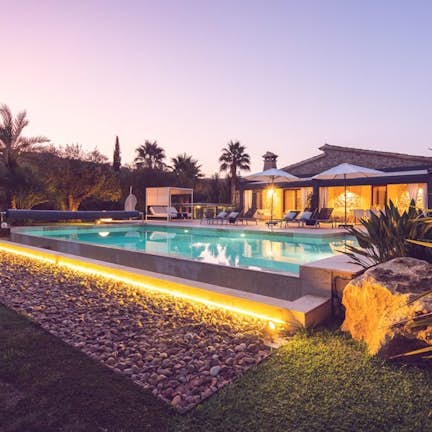 Luxurious house with illuminated swimming pool at twilight, surrounded by landscaped garden and palm trees.