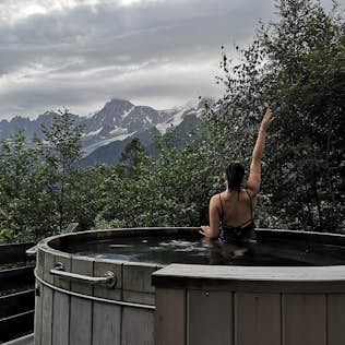 A person in a swimsuit raises an arm while sitting in an outdoor hot tub, mountains and cloudy skies in the background.