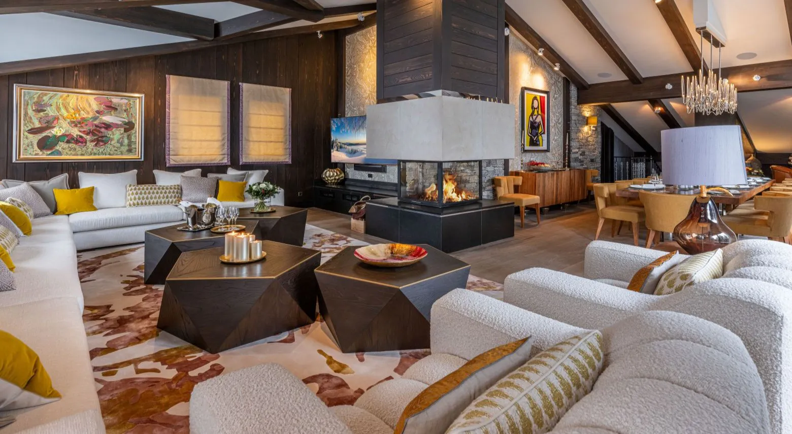 Chalet Coquelicot Spacious lodge living room with plush white sofas, geometric tables, central glass fireplace, beamed ceiling and dining area.
