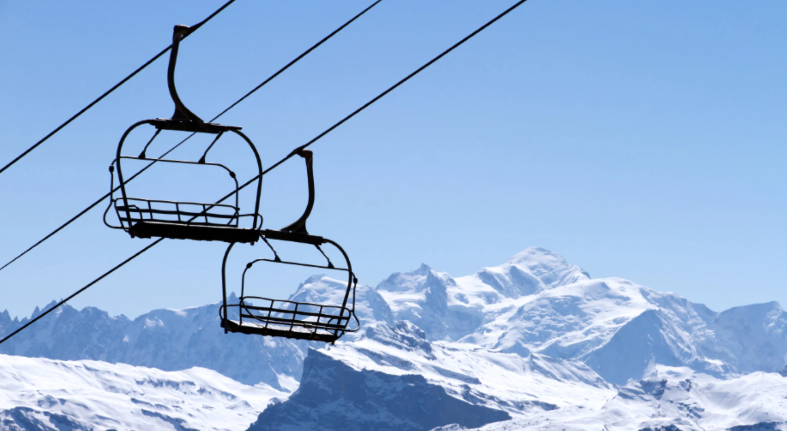 French Ski Resorts Near Geneva | Emerald Stay Two empty ski lift chairs suspended on cables against a backdrop of snowy mountain peaks under a clear blue sky.