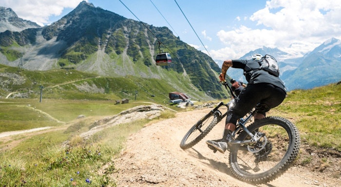 Verbier - Mountain Bike Mountain biker navigating a dirt trail with scenic mountains and a cable car in the background under a blue sky.