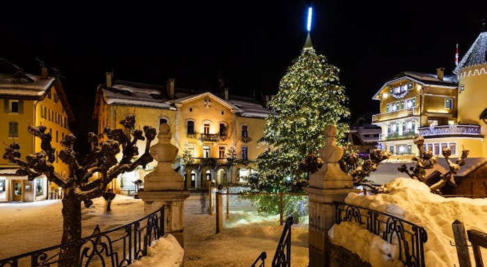 Snowy village square at night with a large, decorated Christmas tree, surrounded by illuminated buildings and snow-covered trees.