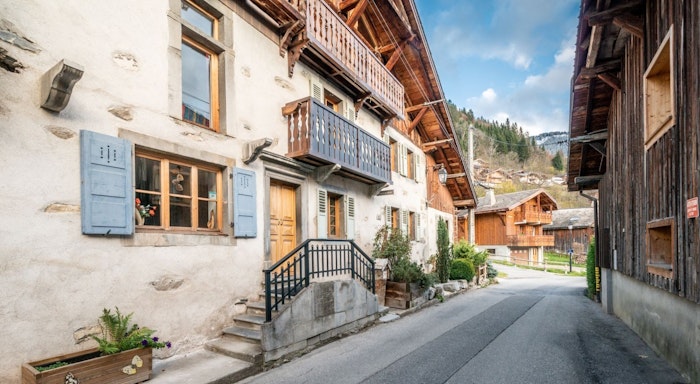 La ferme exterior Charming alpine village street with rustic wooden houses, stone walls, blue shutters, and a mountainous backdrop under a clear sky.