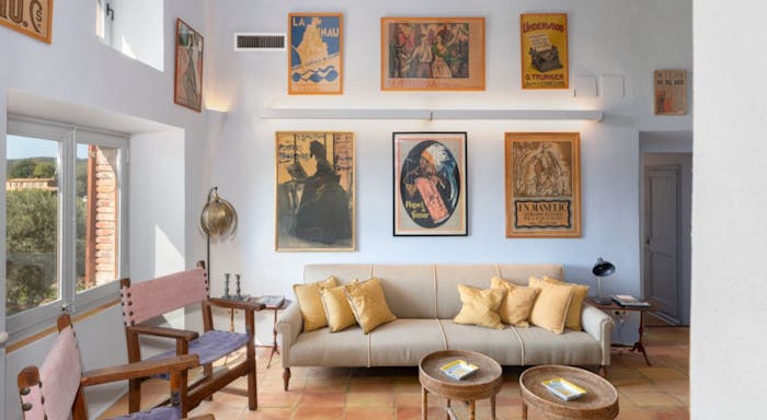 Finca Mas Gotas Interior Bright living room with a beige sofa, yellow pillows, two wooden chairs, round tables, and vintage posters on white walls. Large window on the left.