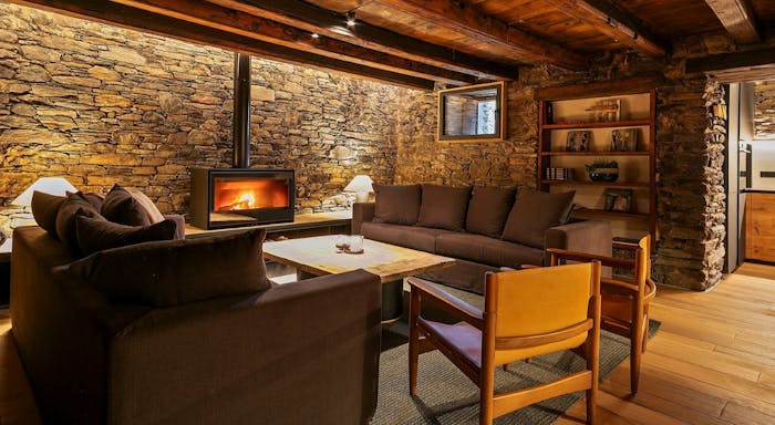 Mas de la costa interior Cozy living room with stone walls, wooden ceiling, and a fireplace. Brown sofas and a wooden chair surround a central coffee table.