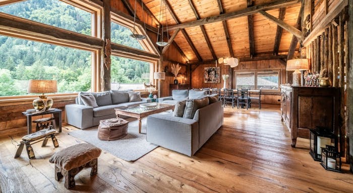 Le soli interior Spacious rustic living room with wooden beams, large windows, gray sofas, and mountain views, featuring cozy decor and natural light.