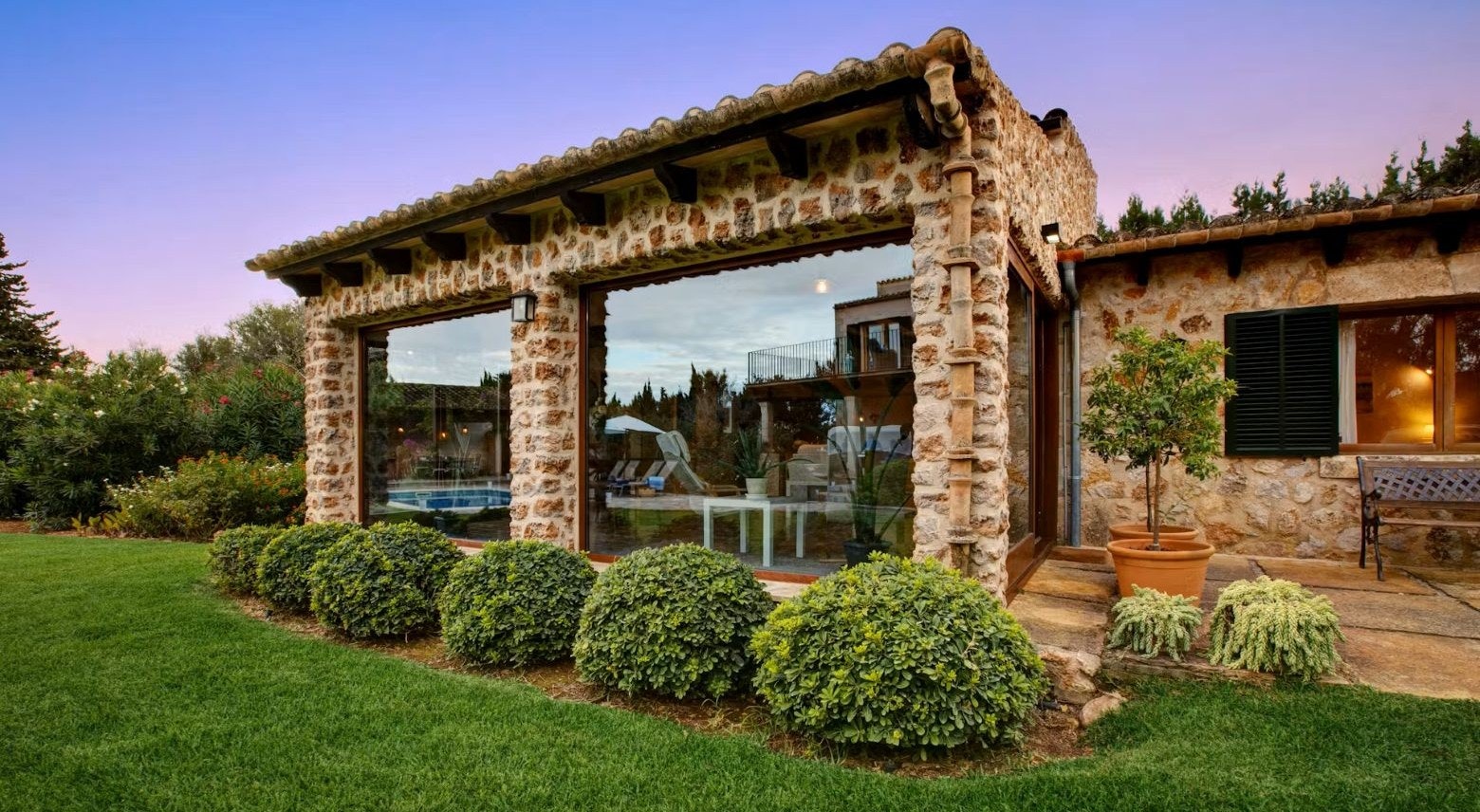 Neil Nightingale: An Award Winning Filmmaker, Owner and Partner A rustic stone house with large windows, surrounded by a well-manicured lawn and shrubbery, near sunset.