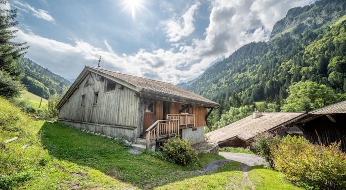 Le soli exterior A rustic wooden cabin nestled in a lush green mountain landscape under a bright, sunny sky with scattered clouds.