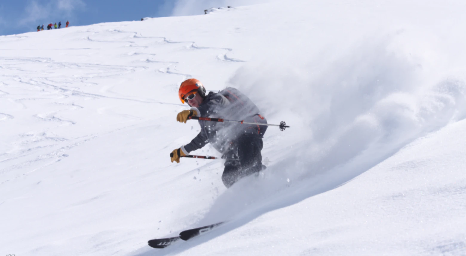 French Ski Resorts Near Geneva | Emerald Stay A skier wearing an orange helmet and black jacket skis downhill through fresh powder on a sunny day, leaving a trail of snow behind.