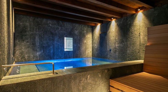 Mas de la costa spa Indoor pool with blue lighting, surrounded by dark stone walls and a wooden ceiling. A wooden bench is on the right side.