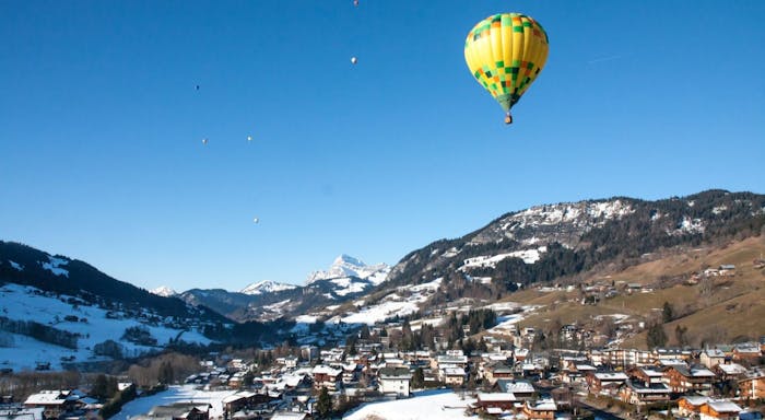 A yellow and green hot air balloon floats over a snowy mountain village under a clear blue sky.