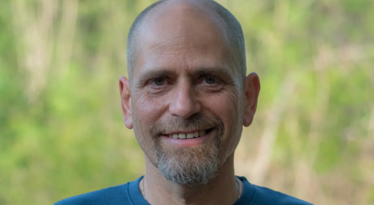 Aviram Rozin, Emerald Stay’s Impact Collaborator A man with a shaved head and short beard smiles at the camera, wearing a blue shirt. The background is blurred greenery.