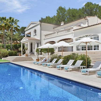 Villa Tranquilo Mallorca Emerald Stay Luxurious villa with a large pool, sun loungers, and umbrellas, surrounded by lush greenery and palm trees under a clear blue sky.