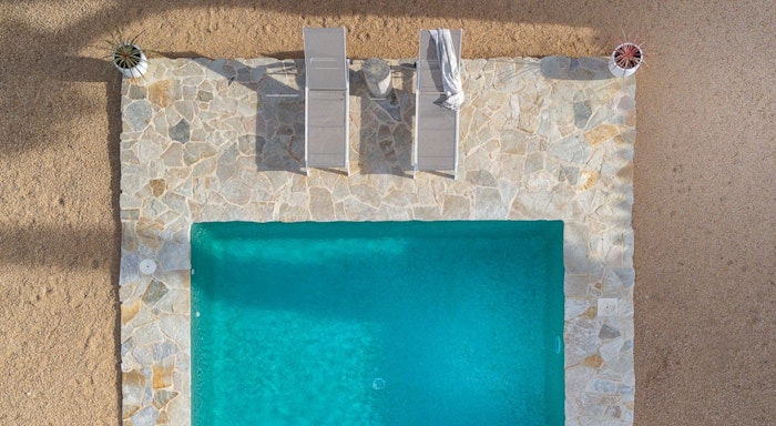 La Capella Aerial view of a rectangular swimming pool with two lounge chairs and a towel on a stone-tiled deck, surrounded by sandy terrain.