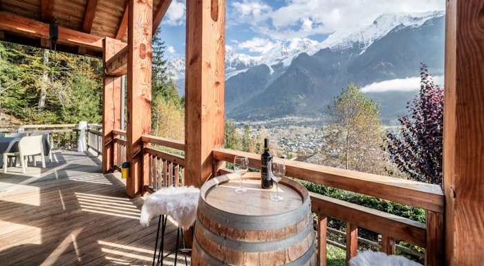 Chalet Ibex Mountain chalet balcony with wooden beams, a wine barrel table, two glasses, and scenic view of snow-capped peaks and a valley below.