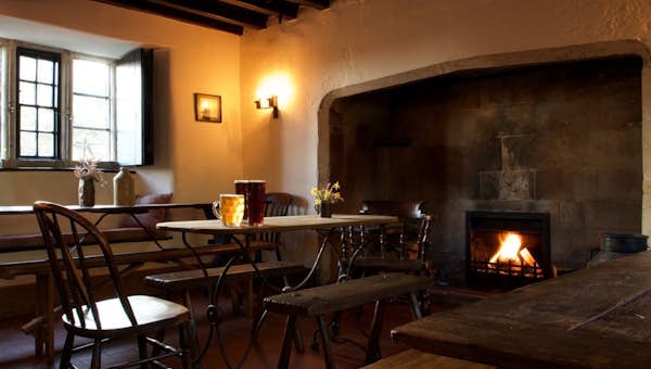 Cozy room with wooden furniture, a wooden table, chairs, benches, a lit fireplace, and a window, creating a warm and inviting atmosphere.