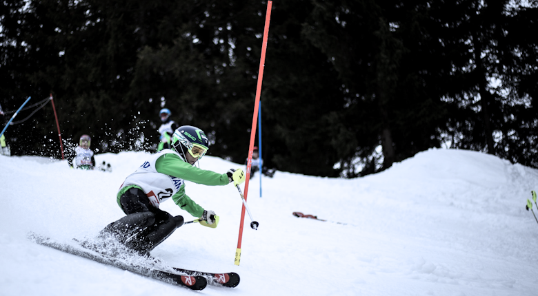 A skier in a green and white outfit navigates a slalom course, leaning through a gate in snowy terrain, with other skiers in the background.
