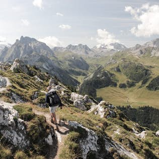 A person with a backpack hiking along a narrow trail in a scenic mountainous region, with rugged peaks and green valleys stretching into the distance.