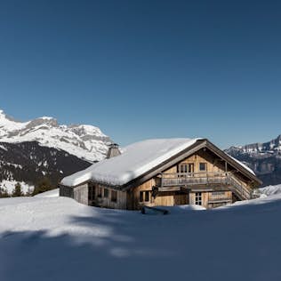 A wooden chalet covered in snow with the swiss alps in the background under a clear blue sky.