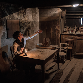 A person in period clothing sits at a wooden table in a dimly lit rustic room, pointing at something while holding a candle.