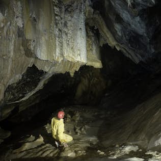 A person in a yellow jacket and red helmet exploring a dark, spacious cave with large stalactites and glossy cave walls.