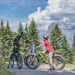 Two mountain bikers wearing helmets high-five on a forest trail with scenic mountain views in the background.