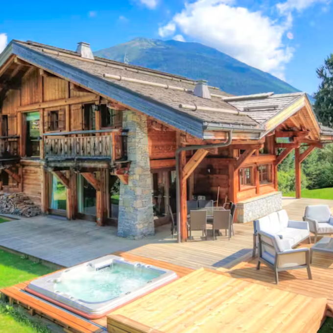 Service de conciergerie à Megève  Conciergerie Megève A spacious wooden cabin with large windows, a patio, a hot tub, and a small pool, set against a mountain backdrop and surrounded by greenery under a blue, cloudy sky.