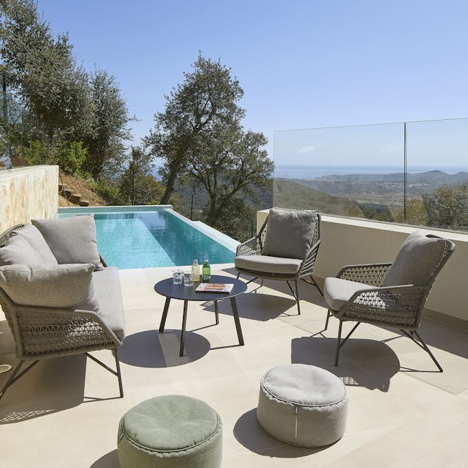 Casa Pere in Costa Brava Outdoor patio with wicker chairs, a small table, and a pool overlooking a scenic landscape with trees and distant water view.