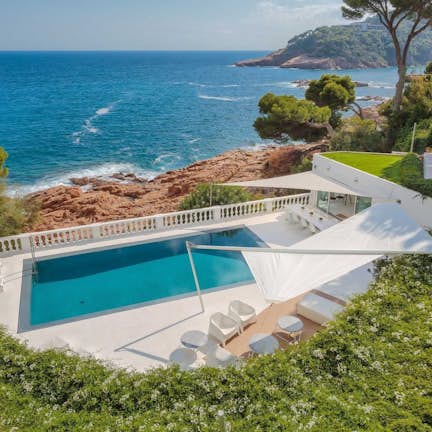 Luxurious seaside villa with a private pool, white lounge chairs under a canopy, surrounded by lush greenery, overlooking a rocky coastline and blue sea.
