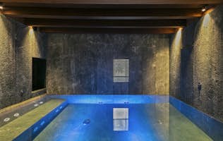 Tarter-Canillo - 4 Andorre location - Chalet Mas de la Costa - Indoor pool with blue lighting, surrounded by stone walls and wooden ceiling beams. There's a wooden bench on the right.