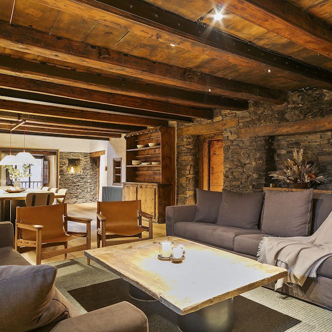 Tarter-Canillo - 1 Andorre location - Chalet Mas de la Costa - Cozy living room with a stone accent wall, dark sofas, a wooden ceiling, and a lit fireplace. Two wooden chairs are around a coffee table. Warm ambient lighting enhances the room.