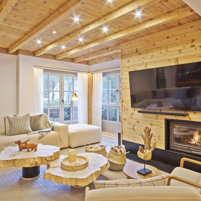 Val de Ruda - 1 Baqueira Beret accommodation - Apartment Aliso - Modern living room with light wood ceiling beams, large windows, a gray sectional sofa, a wooden coffee table, poufs, and a fireplace with a TV mounted next to it. Scenic mountains are visible outside.
