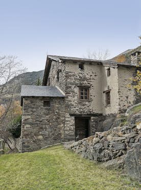 Tarter-Canillo - 5 Andorre location - Chalet Mas de la Costa - A rustic stone building on a grassy slope with bare trees and a wooden fence in the foreground. Mountains are visible in the background under a clear sky.