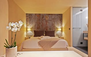 Tarter-Canillo - 2 Andorre location - Chalet Mas de la Costa - Modern room featuring a double bed with brown pillows and beige throw, wooden wall, soft lighting, and a vase with white orchids on a light-colored table.